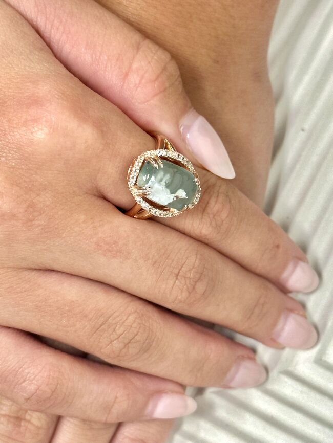 A close-up of a hand wearing a gold ring with a round green shell inlay and a halo of small clear stones, set against a light textured background.
