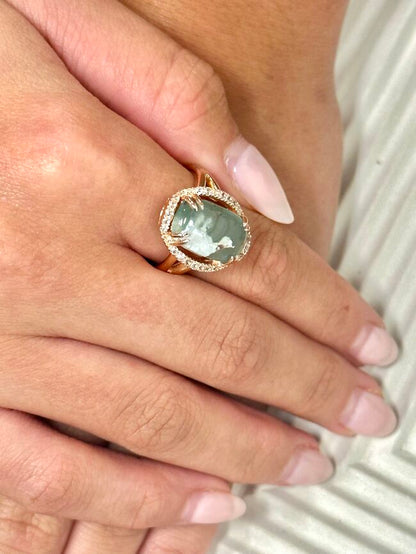 A close-up of a hand wearing a gold ring with a round green shell inlay and a halo of small clear stones, set against a light textured background.